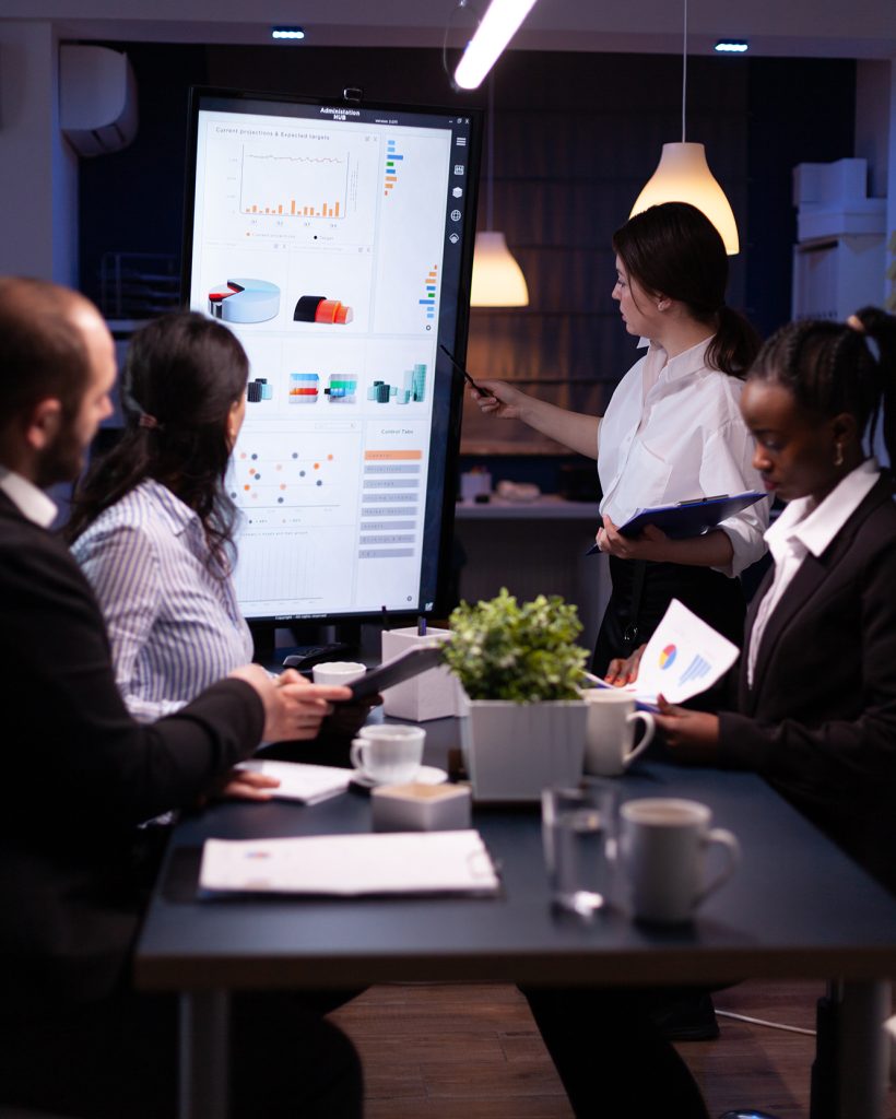 Diverse multiethnic business teamwork overworking in office meeting room analyzing financial graphs late at night. Coworkers brainstorming project ideas solving company problem.